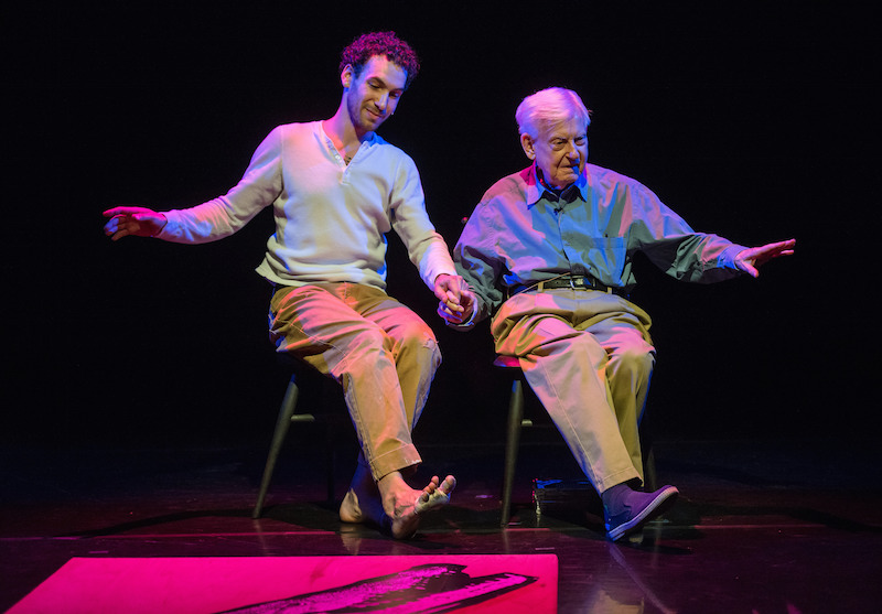 Pepper and David sit on small wooden stools. They stick out their right heels and hold hands.
